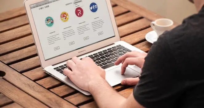 Obrazek przedstawiający osobę korzystającą z laptopa Macbook Air marki Apple, przeglądającą strony internetowe. Laptop znajduje się na drewnianym stoliku, tuż obok kubka z kawą. Osoba ma na sobie czarną koszulkę.